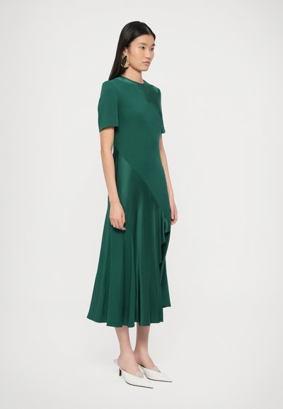 Zelené saténové šaty s krátkými rukávy, kulatým výstřihem a asymetrickým lemem. Kombinovány s bílými mulemi s špičatou špičkou. Hladká textura.