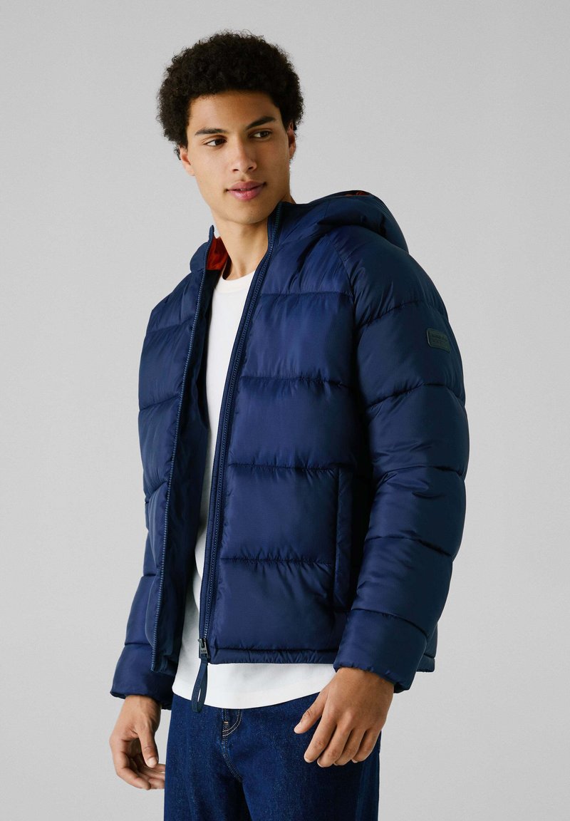 Marineblauwe pufferjack met een capuchon, voorzien van een ritssluitingen, gequilt textuur en contrasterende rode voering. Draagt over een wit shirt.