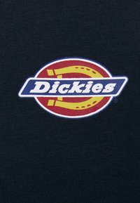 Black fabric features a circular logo with a red background, yellow accents, and white lettering spelling "Dickies" in a bold font.