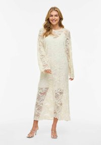 Femme portant une longue robe en dentelle crème transparente avec des motifs floraux et des sandales à talons hauts argentées, souriante devant un fond uni.