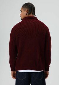 Man met gevlochten haar, gekleed in een donkerrode geribbelde trui en donkere spijkerbroek, gezien van achteren tegen een effen lichte achtergrond.