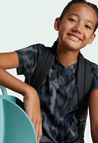 Maglietta sportiva nera e grigia con stampa leopardata, maniche corte e scollatura rotonda, arricchita da una tracolla nera dello zaino e un oggetto color teal.