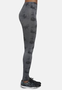 Szare legginsy z czarnym abstrakcyjnym wzorem, wysoki pas, elastyczny materiał i dopasowany krój. Noszone z czarnymi butami sportowymi.