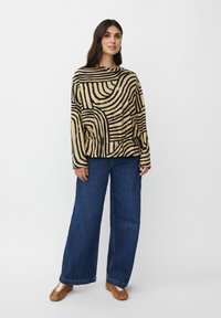 Maglietta a maniche lunghe beige con strisce ondulate nere, abbinata a jeans a gamba larga blu. Il modello indossa scarpe marroni e accessori minimal.