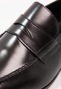 Gros plan sur une chaussure mocassin en cuir noir poli avec des coutures détaillées et une bride classique penny sur le dessus.