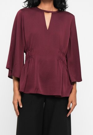 Vrouw draagt een bordeaux blouse met lange mouwen, een kleine voorsplit in de halslijn en geplooide zijkanten, gecombineerd met een zwarte broek.