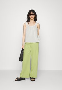 Top branco sem mangas com decote franzido, combinado com calças largas verde claras. Os acessórios incluem uma bolsa tecida preta e óculos de sol escuros.