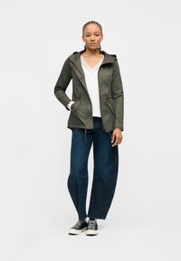 Jeune femme debout portant une veste à capuche vert olive, un haut blanc orné de dentelle, un jean large bleu foncé et des baskets noires sur fond blanc.