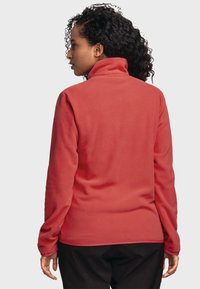 Roter Fleece-Pullover mit hohem Kragen und langen Ärmeln. Besitzt eine glatte Textur und einen dezenten Saum, getragen mit schwarzen Hosen.