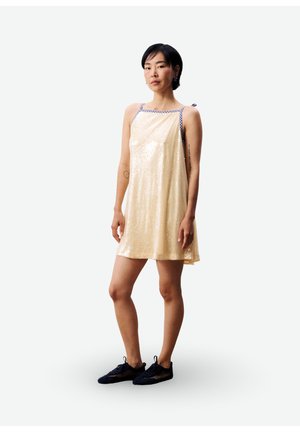 Jonge vrouw staand, gekleed in een lichtgele jurk met pailletten, geruite bandjes en zwarte sneakers, op een witte achtergrond.