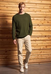 Un pull côtelé vert foncé, un pantalon beige et des baskets blanches. L'arrière-plan présente des planches de bois, créant un contraste de texture naturelle.