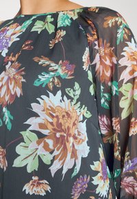 Blomstret bluse med sort gennemsigtigt stof, prydet med farverige blomster og blade i nuancer af grøn, lilla og orange. Blød tekstur, løs pasform.