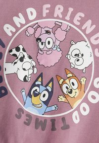 Una tela rosa presenta un diseño circular con cinco personajes de dibujos animados: un perro azul, un perro naranja, una caniche rosa, un perro blanco y negro, y una oveja blanca.
