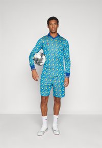 Blaues gemustertes Fußballtrikot mit langen Ärmeln und Shorts, das mit Rauten in Grün und Gelb verziert ist, kombiniert mit einem schwarz-weißen Fußball.
