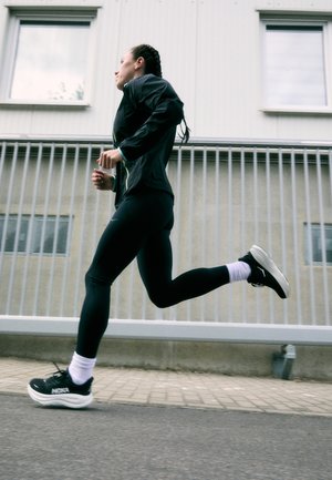 Jaqueta atlética preta com detalhes em neon, leggings pretas e tênis de corrida pretos com solas brancas, contra um fundo cinza claro.