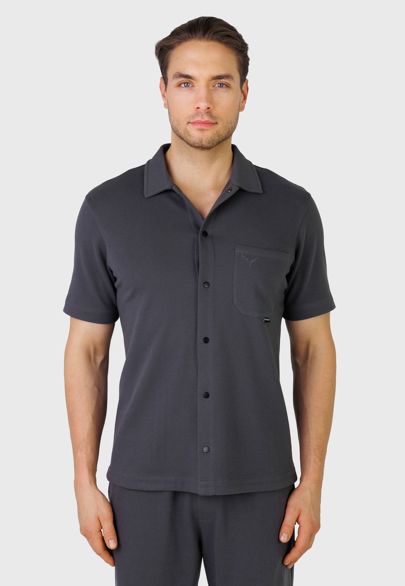 Homme portant une chemise à manches courtes gris foncé avec une poche poitrine, debout devant un fond clair uni.