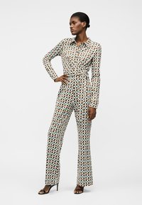 Γυναίκα όρθια με το χέρι στη μέση, φορώντας ολόσωμη φόρμα με γεωμετρικό μοτίβο και μαύρες ανοιχτές γόβες, σε λευκό φόντο.