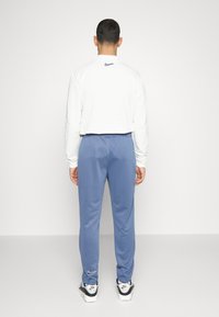 Weißes Langarmhemd und blaue Sweatpants, beide mit lockerer Passform. Nike-Logo auf der Rückseite des Hemdes und unten links an der Hose.