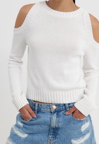 Biały dzianinowy sweter z odsłoniętymi ramionami, prążkowanym dekoltem i mankietami, zestawiony z postrzępionymi szortami z denimu.