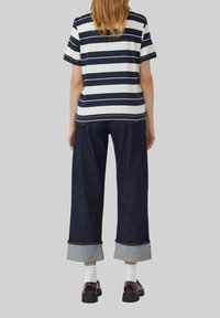 Persoon staat met de rug naar de kijker, gekleed in een marineblauw en wit gestreept T-shirt met korte mouwen, brede donkere jeans met omslagen, witte sokken en donkere schoenen.