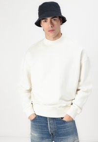 Sweatshirt blanc à col rond en coton, doté d'une coupe décontractée et de poignets côtelés. Associé à un jean en denim bleu déchiré et un chapeau seau noir.