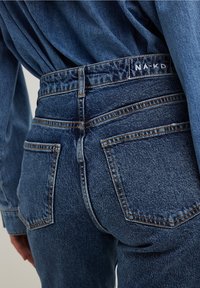 Jeans in denim blu scuro con vita alta, ampie tasche posteriori e dettagli di cucitura. Etichetta "NA-KD" visibile sulla vita posteriore.