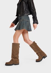Stivali marroni in suede al ginocchio con gambale largo, suola in gomma nera, zip laterale e punta arrotondata, abbinati a una minigonna di jeans e una giacca di pelle.