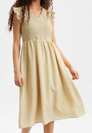 Kobieta ubrana w żółtą i białą sukienkę w kratę bez rękawów z falbankami na ramionach i przyszytym karczkiem, trzymająca spódnicę jedną ręką.