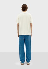 Camiseta de hombre de corte holgado en color crema y de mangas cortas, con detalle en la parte trasera del cuello, combinada con unos vaqueros de pierna ancha azul, vista desde la parte trasera.