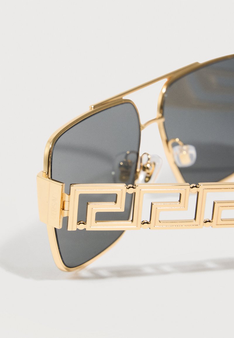 Gold-framed sunglasses with dark lenses featuring a geometric Greek key pattern detail on the arms against a white background.