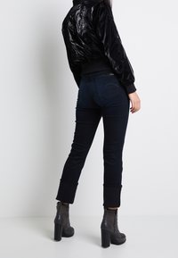 Blouson aviateur noir à texture brillante, associé à un jean en denim foncé avec une jambe fuselée et un ourlet retroussé, complété par des bottines à talons.