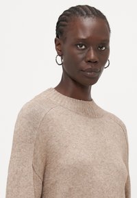 Maglione beige a maglia con scollatura rotonda, dettagli a costine sul collo e design senza cuciture alle spalle, indossato da una modella con capelli intrecciati.