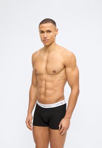 Μυώδης άνδρας με κοντά μαλλιά που φοράει μαύρα boxer Tommy Hilfiger με λευκή μέση, στέκεται μπροστά σε απλό ανοιχτό φόντο.