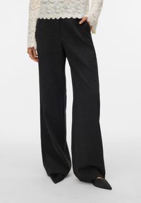 Wijd uitlopende broek in donkergrijze stof met een gladde textuur en een klassieke snit. Gecombineerd met zwarte puntige schoenen voor een strakke uitstraling.