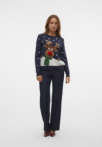 Navy sweater featuring a reindeer with antlers, red nose, green scarf, and snowflake pattern. Paired with dark wide-leg jeans and red shoes.