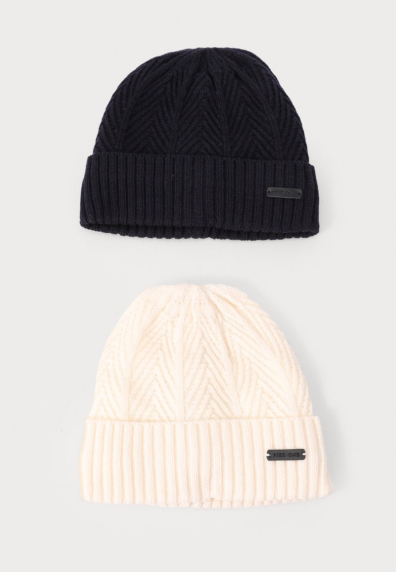 Two knitted beanies: one dark navy with a ribbed cuff and zigzag texture; one cream with similar texture and ribbed cuff. Both feature small tags.