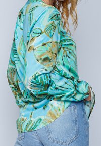 Blusa a maniche lunghe in turchese con stampa floreale, realizzata in tessuto morbido e liscio, con una vestibilità rilassata e un orlo curvo, abbinata a jeans chiari.