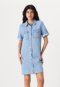Lys blå denimkjole med korte ærmer, knappefront og to brystlommer. Glatt tekstur, uformelt design og knelange snitt.