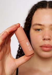 Rosa sylinderformet leppeprodukt som holdes mellom fingrene, med en matt finish, hvit tekst og subtile merkevareelementer.