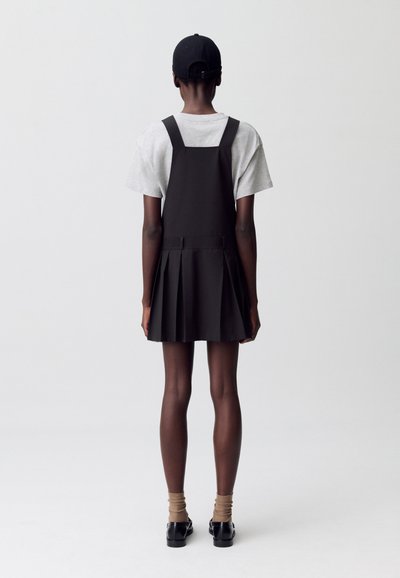 Una persona está de pie de espaldas, vistiendo una camiseta gris y un vestido negro con una falda plisada, combinado con calcetines marrones y zapatos negros.