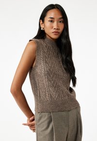 Ärmelloser, hochgeschlossener, brauner Strickpullover mit strukturierten Kabelmustern und geripptem Saum, kombiniert mit grauen, maßgeschneiderten Hosen.