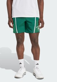 Žalios sportinės šortai, pagaminti iš lengvos medžiagos, su baltais šoninėmis juostelėmis, logotipu šone ir klasikinio sportinio dizaino.