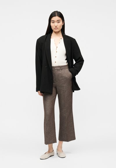 Femme portant un blazer noir oversized, un haut blanc boutonné, un pantalon large marron et des ballerines beige, debout avec une main dans la poche.