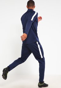 Marinblå träningsdräkt med vita sidostreck, tillverkad av ett lätt material. Fitted design med ribbad mudd och krage. Svarta skor som accent.