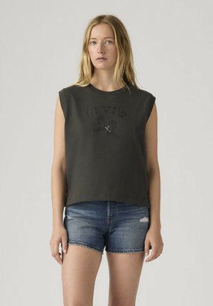 Mujer con camiseta negra sin mangas con diseño gráfico y pantalones cortos de mezclilla azul, de pie contra un fondo liso gris claro.