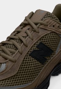 Καφέ αθλητικό παπούτσι από διχτυωτό ύφασμα και σουέτ με χοντρά κορδόνια και μαύρο λογότυπο "N" στο πλάι, τοποθετημένο σε απλό λευκό φόντο.