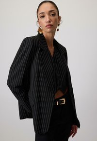 Blazer negru cu dungi subțiri, cu rever larg, asortat cu o cămașă neagră cu nasturi și o curea neagră cu cataramă aurie. Material neted.
