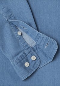 Dettaglio di una camicia di jeans blu chiaro con colletto a punta, due bottoni bianchi e un dettaglio cucito sui polsini con asole per bottoni.