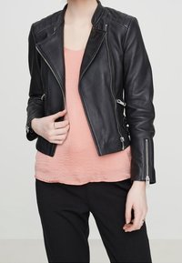 Person som bär en svart läderjacka med dragkedjeslut över en persikofärgad texturerad blus och svarta byxor, som justerar jackan med ena handen.