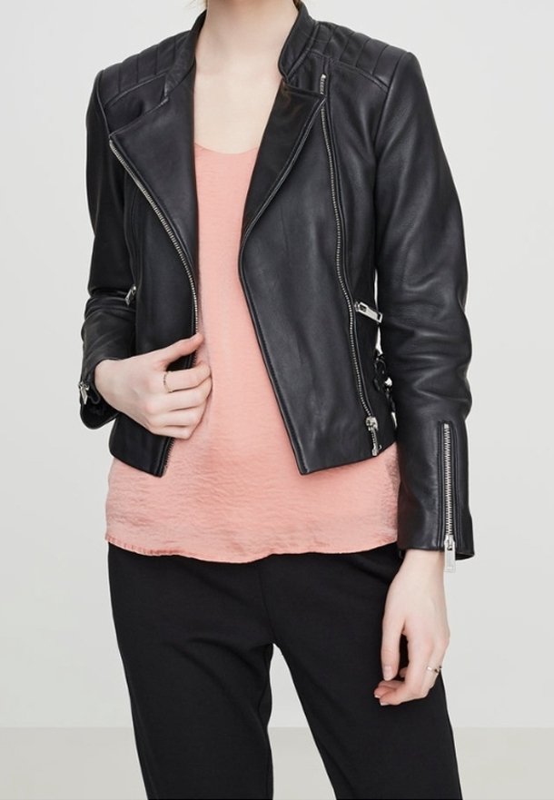 Person som bär en svart läderjacka med dragkedjeslut över en persikofärgad texturerad blus och svarta byxor, som justerar jackan med ena handen.